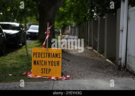 Bright yellow pedestrians watch your step sign, featuring black text, near a stretch of unpaved footpath, under shady trees in a leafy green suburb Stockfoto