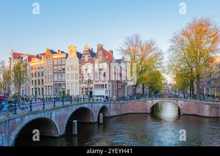 Goldene Blätter flattern in der frischen Herbstbrise, während Radfahrer die friedlichen Brücken in Amsterdam Keizersgracht überqueren, umgeben von bezaubernden Kanalhäusern, die die Schönheit und Ruhe der Jahreszeit widerspiegeln. Stockfoto