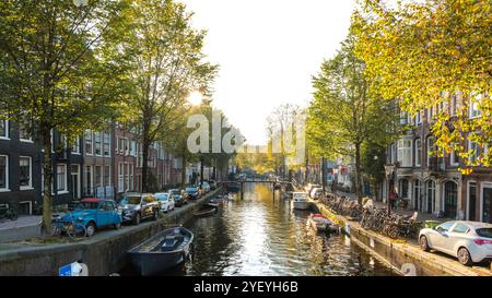 Goldene Blätter flattern in der frischen Herbstbrise, während die Sonne über Amsterdams ruhigen Kanälen untergeht. Die Boote gleiten sanft vorbei, reflektieren die warmen Farben der Natur und laden zu Spaziergängen auf von Bäumen gesäumten Pfaden ein. Stockfoto