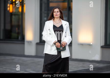 Stilvolle Frau in Übergröße steht draußen im urbanen Ambiente der Stadt und trägt ein modisches schwarzes Kleid mit weißem zweireihigem Blazer-Outfit. Modell mit Stockfoto