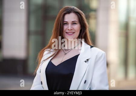 Porträt lächelnde Frau in Übergröße steht draußen im urbanen Ambiente der Stadt und trägt ein modisches schwarzes Kleid mit weißem zweireihigem Blazer-Outfit. M Stockfoto
