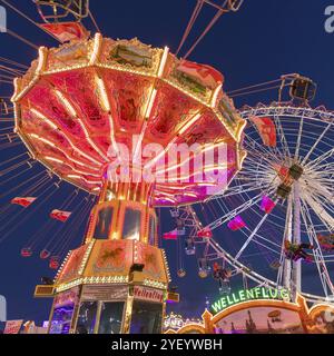 Ein Jahrmarkt in der Abenddämmerung mit beleuchtetem Kettenkarussell und Riesenrad, Europa Rad, Fahrten, Wellenflug, Cannstatter Wasen, Volksfest Bad Cannstatt, S Stockfoto