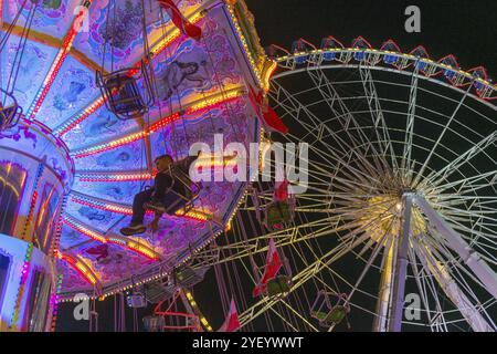 Ein Jahrmarkt in der Abenddämmerung mit beleuchtetem Kettenkarussell und Riesenrad, Europa Rad, Fahrten, Wellenflug, Cannstatter Wasen, Volksfest Bad Cannstatt, S Stockfoto