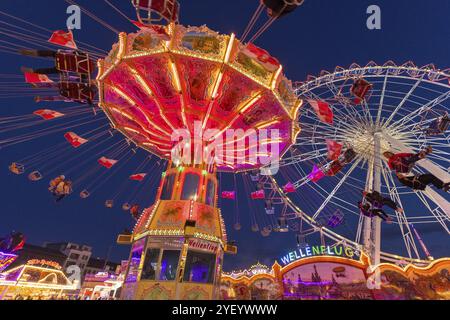 Ein Jahrmarkt in der Abenddämmerung mit beleuchtetem Kettenkarussell und Riesenrad, Europa Rad, Fahrten, Wellenflug, Cannstatter Wasen, Volksfest Bad Cannstatt, S Stockfoto