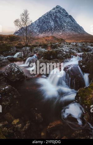 Ein Wasserfall fließt unter dem schneebedeckten Berg Buachaille Etive Mòr in Glencoe, Schottland, und unterstreicht die dramatische Schönheit der Scottis Stockfoto