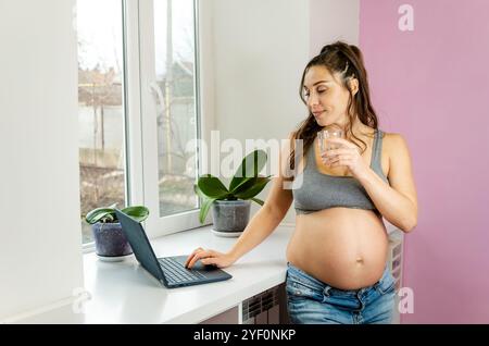 Eine glühende schwangere Frau bleibt während der Arbeit an ihrem Laptop hydratisiert Stockfoto