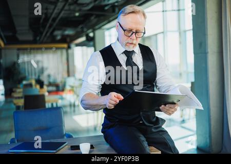 Ein grauhaariger bärtiger Mann in Brille und weißem Hemd sieht sich Dokumente an. Ein erwachsener Mann sitzt am Schreibtisch im Büro. Stockfoto