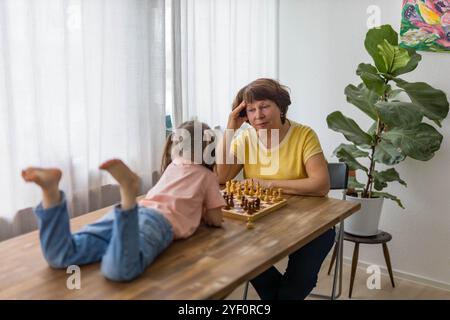 Ein fröhliches Mädchen spielt Schach mit ihrer Großmutter am Tisch und schafft wertvolle Familienerinnerungen. Hochwertige Fotos Stockfoto