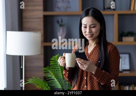 Asiatische Frau zu Hause lächelt, während sie ihr Smartphone benutzt und Kaffee trinkt. Lässig und lässig ist sie in brauner Bluse mit Punktmuster und genießt moderne Kommunikation in elegantem Wohnklima. Stockfoto