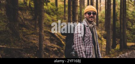 Porträt eines jungen gutaussehenden Mannes mit Rucksack, Sonnenbrille und gelbem Hut, der im Wald in die Kamera lächelt Stockfoto