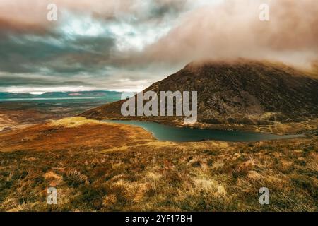 Ein Berghang mit einem blauen See an der Basis, umgeben von trockenem Gras und tiefer Vegetation, mit Wolken, die den Berggipfel verdecken. Stockfoto