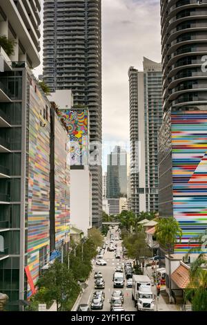 Miami, Florida, USA - 4. Dezember 2023: Verkehr und Fußgänger auf einer Straße in der Innenstadt zwischen Gebäuden mit farbigen Außenwänden. Stockfoto