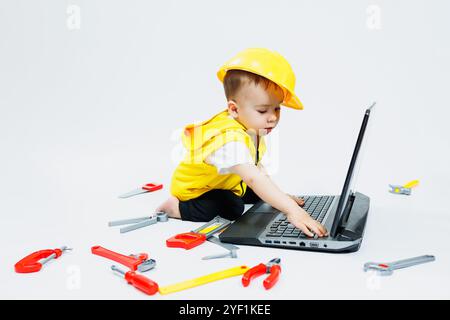 Ein zweijähriger Junge in einer gelben Weste auf weißem Hintergrund sitzt mit einem Laptop und spielt mit Plastikbauwerkzeugen. Spielzeug für kleine Kinder. Stockfoto