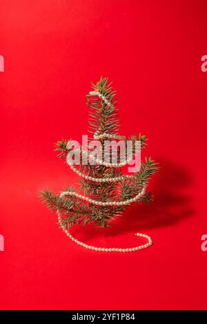 Minimalistischer Weihnachtsbaum aus immergrüner Pflanze und weißen Perlen Halskette auf rotem Hintergrund. Neujahrskonzept. Flache Lagen. Stockfoto