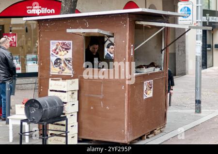 Viladecans. Spanien - 2. November 2024: Kastanien- und Süßkartoffelstand mit einer Verkäuferin und einigen Kisten hinter einer schwarzen Kehrmaschine, die als Stand dient Stockfoto