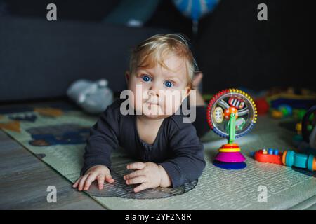 Ein 7 Monate altes Baby krabbelt auf einem Teppich. Das Kind hält den Kopf. Ein Baby lernt, auf dem Boden zu krabbeln. Ein Kind in einem grauen Strampelanzug. Stockfoto