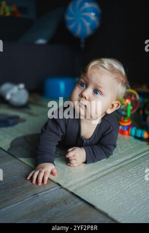 Ein 7 Monate altes Baby krabbelt auf einem Teppich. Das Kind hält den Kopf. Ein Baby lernt, auf dem Boden zu krabbeln. Ein Kind in einem grauen Strampelanzug. Stockfoto