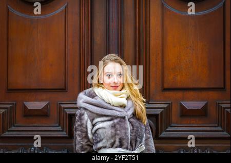 Eine Frau mit blondem Haar und einem warmen Pelzmantel steht vor einer kunstvollen Holztür. Sie trägt einen hellen Schal und schaut direkt auf das Kommen Stockfoto