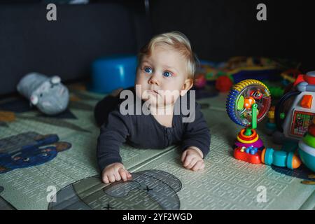 Ein 7 Monate altes Baby krabbelt auf einem Teppich. Das Kind hält den Kopf. Ein Baby lernt, auf dem Boden zu krabbeln. Ein Kind in einem grauen Strampelanzug. Stockfoto