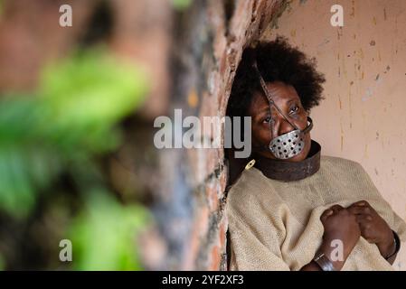 Schwarze Frau gekettet und mit einer eisernen Maske auf dem Gesicht, die gegen eine Ziegelmauer gelehnt ist. Darstellung der Sklaven Anastacia. Sklaverei in Brasilien. Stockfoto