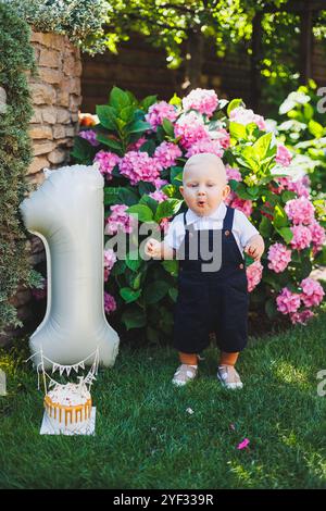 Der erste Geburtstag des Jungen mit einem aufblasbaren Ball. Ein Junge in einem weißen Poloshirt und Overall. Urlaubsbekleidung für Babys. Stockfoto