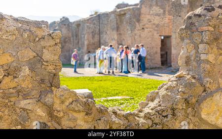 Gruppe von Touristen, die dem Reiseleiter an der archäologischen Stätte von pompeji lauschen. Blick auf verschwommene Touristen, die durch ein Loch in der Mauer auf die Ruinen schauen. Gut Stockfoto