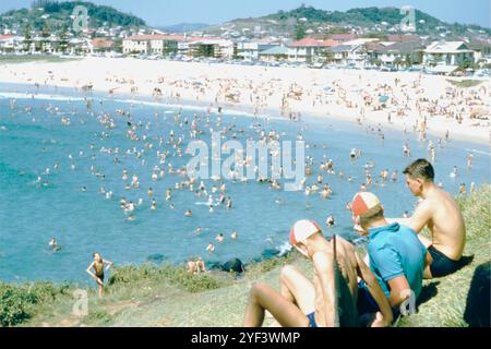 1959 Greenmount Beach, Gold Coast, Queensland, Australien - Eine lebhafte Sommerszene, in der Strandgänger und Surfer die überfüllte Küste von Greenmount Beach überblicken. Das Bild zeigt die ikonischen roten und gelben Kappen, die von Surf-Rettern getragen werden, ein Symbol für Australiens Surf-Rettungsdienste. Im Vordergrund sehen Sie eine Gruppe junger Männer auf dem grasbewachsenen Hügel, mit Blick auf die geschäftige Brandung voller Schwimmer, die das warme Wasser des Ozeans genießen. Dieses historische Foto spiegelt die Strandkultur und den Freizeitstil der australischen Küstenstädte Mitte des 20. Jahrhunderts mit Wohngebäuden wider Stockfoto