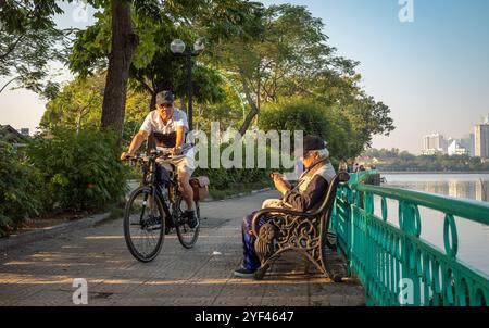 Ein alter Mann sitzt auf einer Bank und schaut auf sein Handy, während ein anderer alter Mann am frühen Morgen am West Lake in Hanoi, Vietnam vorbeifährt. Stockfoto