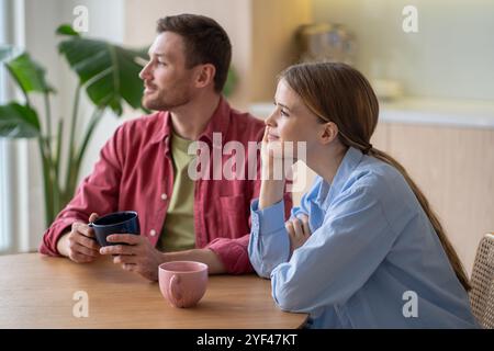 Lächelndes junges Paar, das in die Ferne starrt, während es zusammen Tee und Frühstück in der Küche hatte Stockfoto