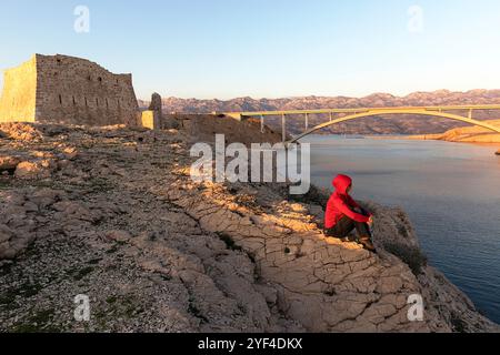 Frau, die bei Sonnenuntergang auf einem Felsen sitzt, an den Ruinen einer antiken Festung Fortica auf der Insel Pag, Dalmatien, Kroatien, pag-Brücke auf der Rückseite Stockfoto