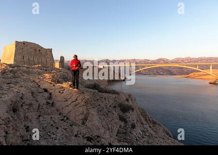 Frau, die bei Sonnenuntergang auf einem Felsen steht, an den Ruinen einer antiken Festung Fortica auf der Insel Pag, Dalmatien, Kroatien, pag-Brücke auf der Rückseite Stockfoto