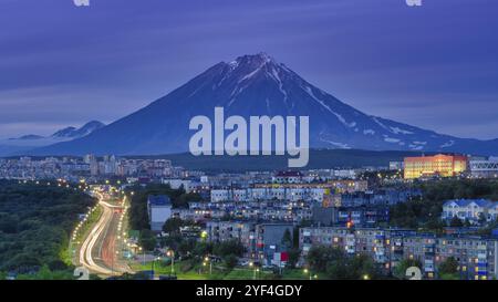 Panorama der Nacht Stadt Petropawlowsk-Kamtschatski auf Hintergrund Kegel des Vulkans, Stadtentwicklung in der Dämmerung, hinterleuchtete Stadtstraße mit dem fahrenden Auto li Stockfoto