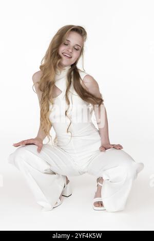 Lächelnde junge Frau mit geschlossenen Augen, sitzende Hockposition, legte beide Hände auf die Hüfte. Verspieltes, sinnliches Blondie-Modell mit langen, welligen Haaren Stockfoto