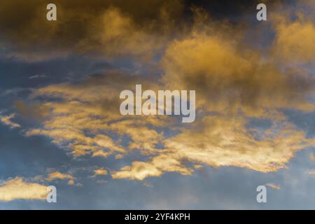 Goldene, flauschige Wolken, beleuchtet von verschwindenden Strahlen bei Sonnenuntergang und dunkle Gewitterwolken, die über den sonnigen blauen Himmel schweben, um das Wetter der Jahreszeit zu ändern. Stunni Stockfoto