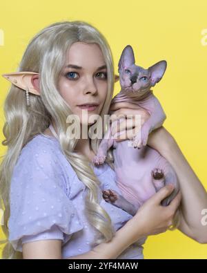 Junge blonde Frau kosplay Elf in blauem Kleid, die Sphinx-Kätzchen in Händen hält und es zeigt. Elfe mit lockigen langen Haaren, Piercings im Ohr, Augen unterschiedlicher Art Stockfoto