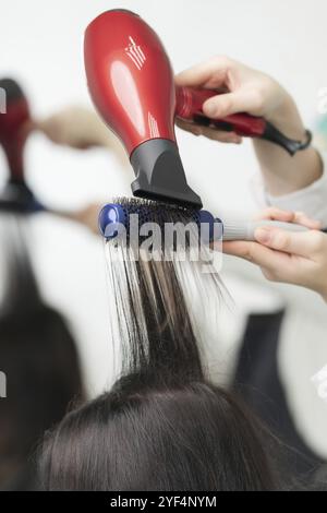 Hände des Friseurs trocknen lange, brünette Haare des Kunden mit rotem Haarfön und blauem Kamm im professionellen Friseursalon Stockfoto