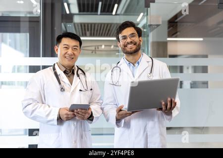 Ärzte stehen im Büro, einer hält Laptop, der andere mit Tablet. Steht für Teamarbeit, professionelle Gesundheitsversorgung und digitale Zusammenarbeit in einem medizinischen Umfeld. Lächelnde, selbstbewusste Profis. Stockfoto