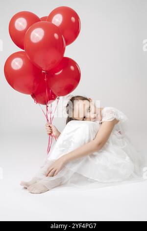 Romantisches Mädchen in weißem Kleid, das viele rote Ballons in der Hand hält, Kopf auf die Knie legt, auf weißem Hintergrund sitzt und in die Kamera blickt. Stu Stockfoto