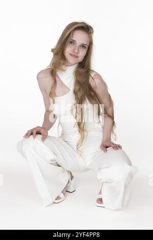 Vorderansicht einer jungen blonden Frau in weißem Jumpsuit und Slingbacks, die in der Hockposition sitzt, beide Hände auf die Knie legte und in die Kamera schaute. Cauca Stockfoto