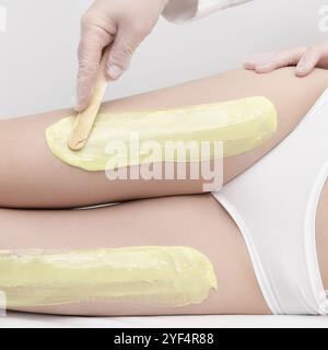 Kosmetikerin in Handschuhen trägt grünes heißes Wachs mit Spachtel auf die schlanken Beine der Frau auf, während die Frau auf der Couch liegt. Enthaarung mit heißem Wachs im Beauty salo Stockfoto