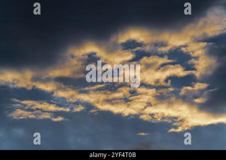 Goldene, flauschige Wolken, beleuchtet von verschwindenden Strahlen bei Sonnenuntergang und dunkle Gewitterwolken, die über den sonnigen blauen Himmel schweben, um das Wetter der Jahreszeit zu ändern. Erstaunlich Stockfoto
