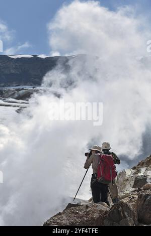 Wandern in Kamtschatka: Zwei Touristen fotografieren und schauen die rauchende (dampfende) Fumarole auf dem aktiven Vulkan Mutnovsky an einem sonnigen Tag. Eurasien Stockfoto