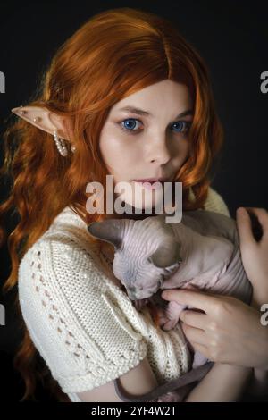 Porträt der jungen Cosplay-Elf-Frau in weißem Strickkleid, die verschlafene Sphinx-Pussycat in den Händen auf schwarzem Hintergrund hält. Blauäugiger Elf mit langer roter Kuh Stockfoto