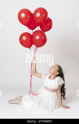 Ein lächelndes Mädchen in einem langen weißen Kleid, das einen großen Haufen roter Ballons in der Hand hält und auf Bälle schaut. Studiofoto eines sitzenden weißen Mädchens Stockfoto