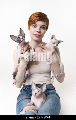 Rothaarige junge Frau mit zwei Sphynx Cat in den Händen und einem Kätzchen, das auf ihren Beinen liegt. Hübsche Frau mit kurzen Haaren in T-Shirt und Jeans suchen Kamera Stockfoto