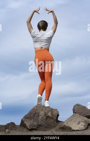 Rückansicht einer Frau, die Yoga praktiziert, auf einem felsigen Berggipfel auf dem Hintergrund des Wolkenhimmels steht. Rückansicht des Körpers einer schlanken Sportlerin. Yoga Stockfoto