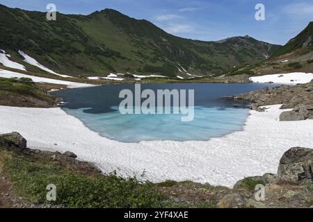 Traumhaft schöne Sommerberglandschaft der Kamchatka Halbinsel: Blauer See, Schnee und Eis entlang der Ufer des Bergsees, grüner Wald auf Hügeln Stockfoto