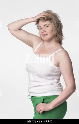 Frau mit praller Figur posiert mit einer Hand auf den Kopf und einer Hand in der Tasche, stehend auf weißem Hintergrund. Authentisches, ausgereiftes Modell mit Blick auf c Stockfoto