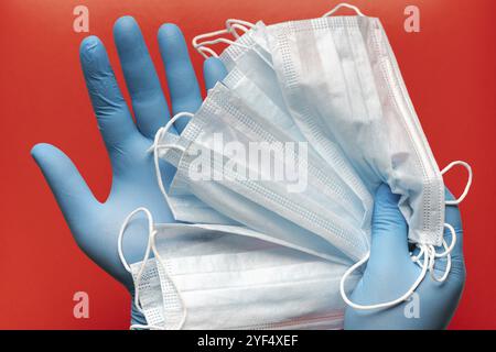 Der Arzt hält die atmungschirurgische Gesichtsmaske in den Händen blaue medizinische Handschuhe auf rotem Hintergrund. Pandemie-Corona-Virus, Versicherungen, durch die Luft übertragbare Krankheiten, SARS Stockfoto