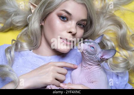 Porträt der jungen Cosplay-Elf-Frau in blauem Kleid, mit luxuriösen lockigen blonden Haaren, Augen in verschiedenen Farben, durchbohrten Ohren. Sie zeigt mit dem Finger auf Sphin Stockfoto